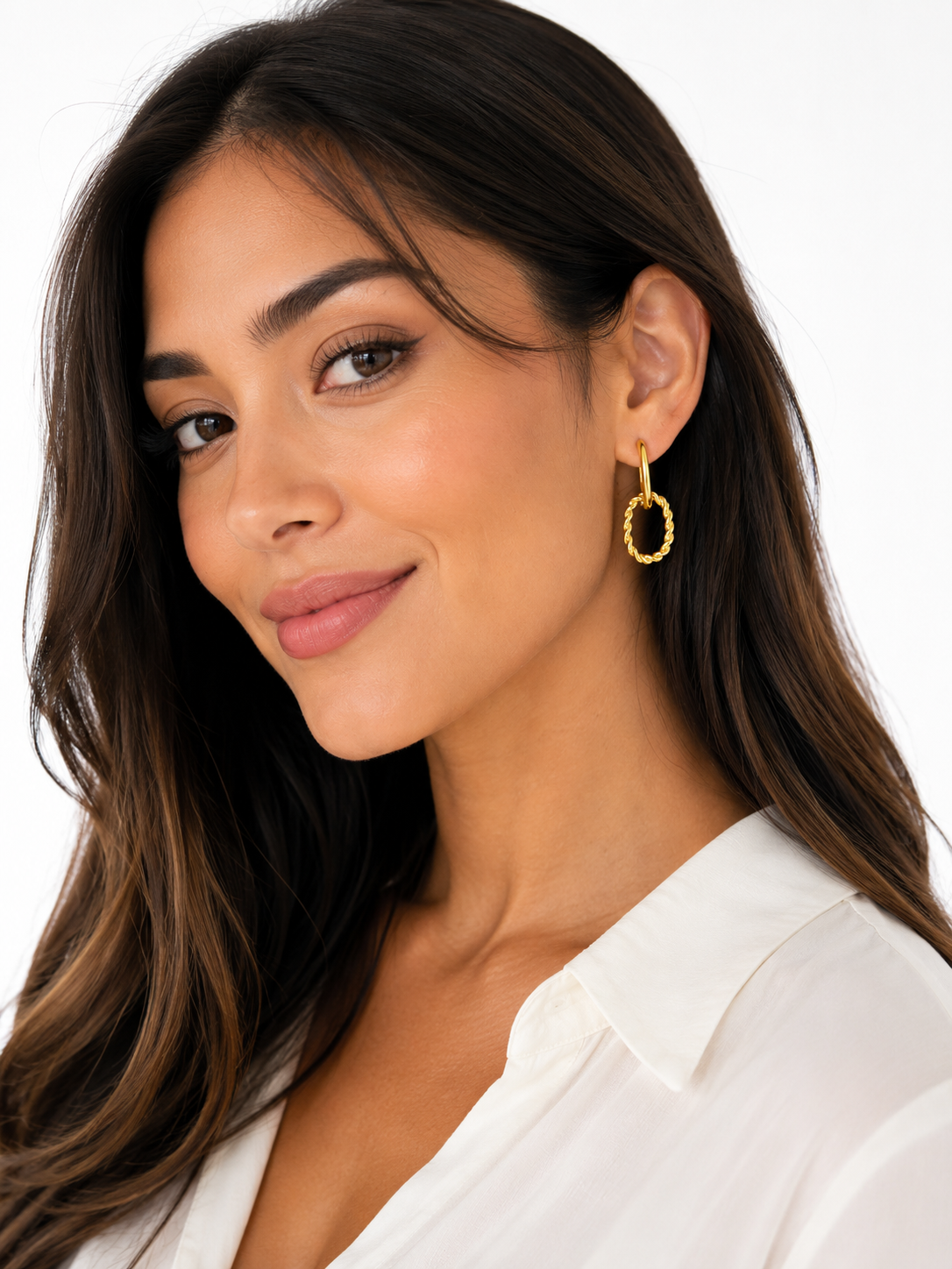 A close-up of a woman wearing a small gold hoop earring with a textured, beaded outer edge, paired with a white collared blouse against a clean white background.