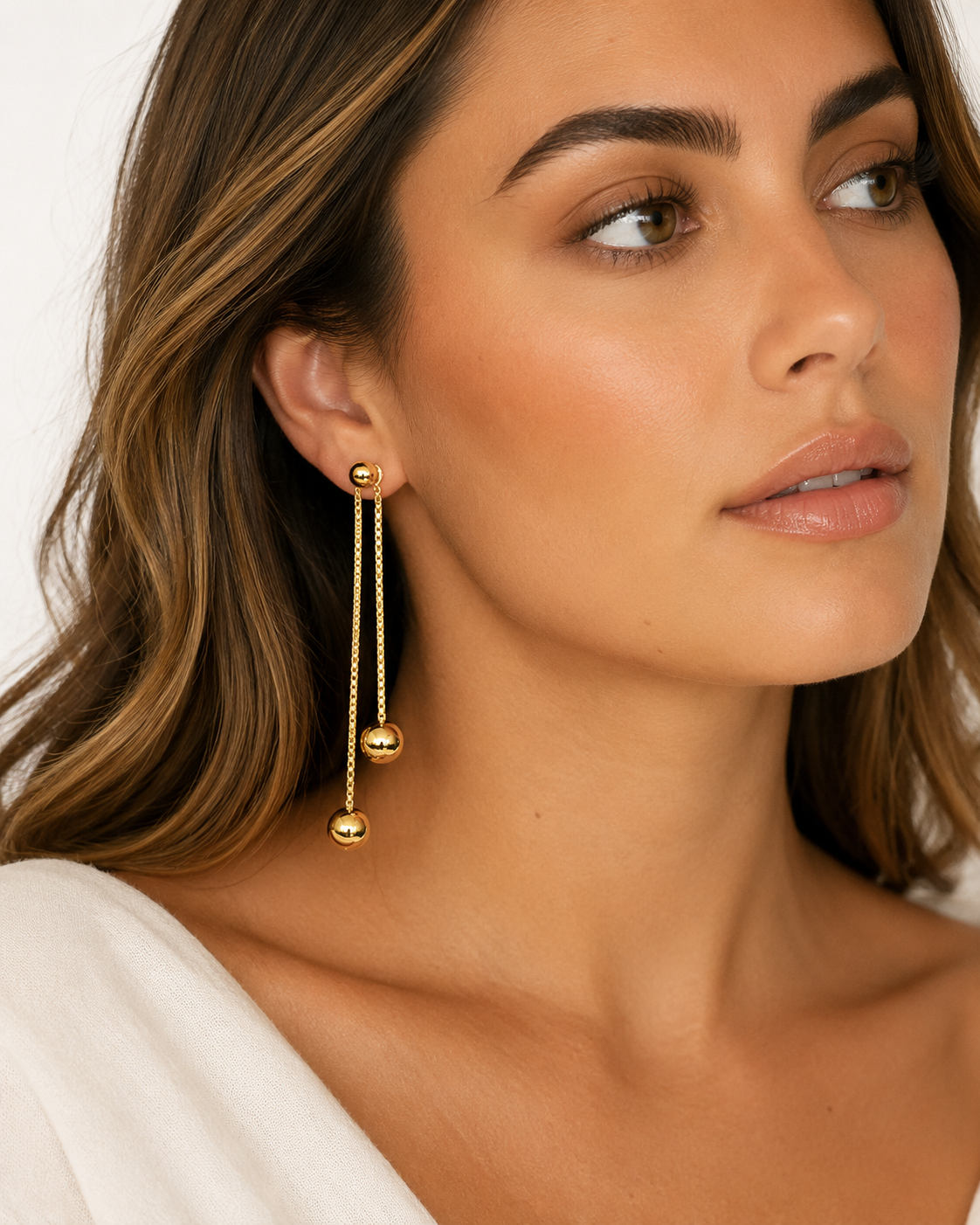 A close-up of a woman wearing a pair of gold dangle earrings with small spherical beads hanging from delicate chains.
