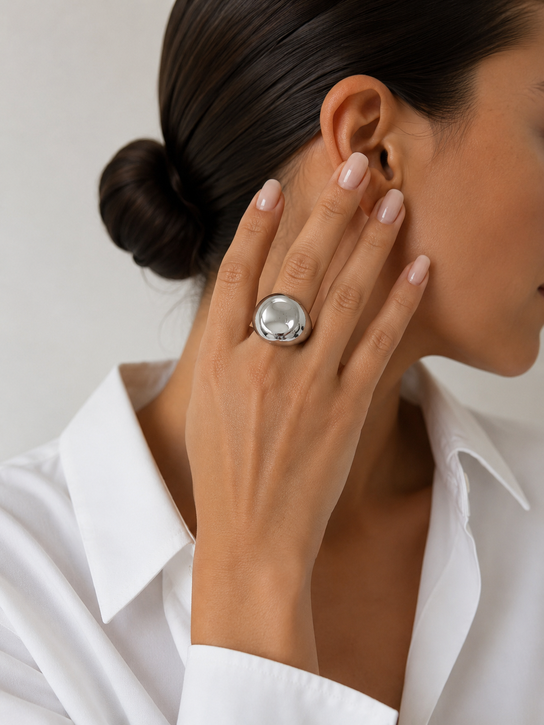 A close-up lifestyle shot of a model wearing a large, chunky silver ring with a polished, dome-like center on her finger. She is wearing a crisp white collared shirt, and her hair is styled in a neat bun against a soft, light gray background.